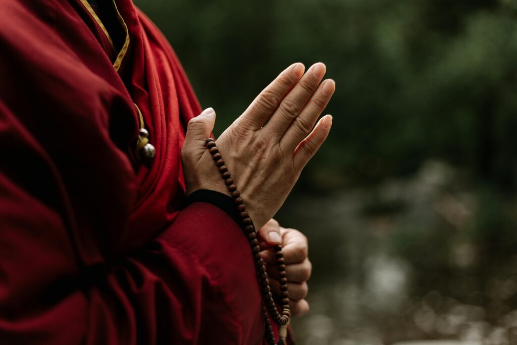 pexels photo 5385913 5385913 Close-up of monk in red robe meditating with prayer beads outdoors.